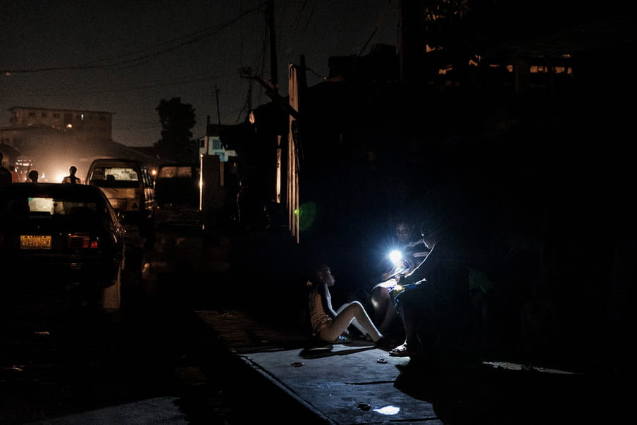 In the darkness of the night, three children are gathered around a source of light hold by one of them. Two are sitting on something unidentified while the third one sits on the ground made of concrete. Some human silhouettes, cars and buildings are visible on the left side of the frame. 