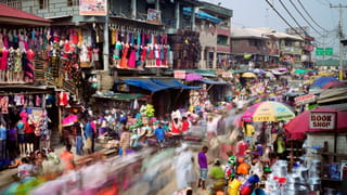Photo of a busy street with a market on it, colourful merchandise hanging everywhere. It's shot with a long shutter speed so the people are blurred