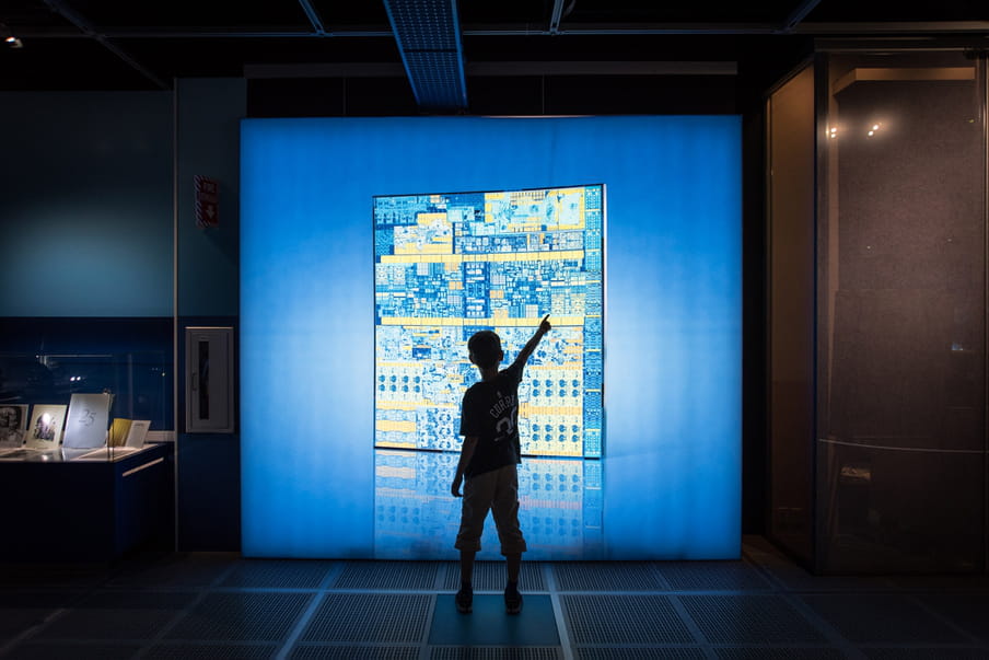 Photograph of a young person’s silhouette standing and pointing to a blue screen wall of what looks like a yellow and blue aerial map of buildings. There are books on display in a glass cabinet to the left