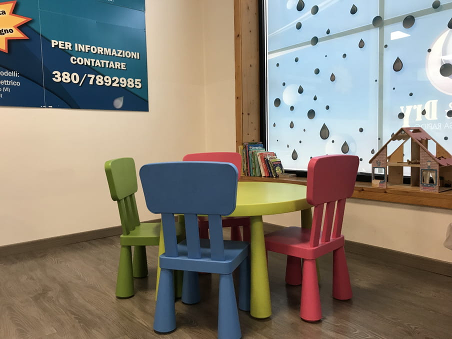 Small plastic chairs in different colours (blue, green and red) are around a small green plastic table. In the background there is a corner with books and a miniature wooden house. 