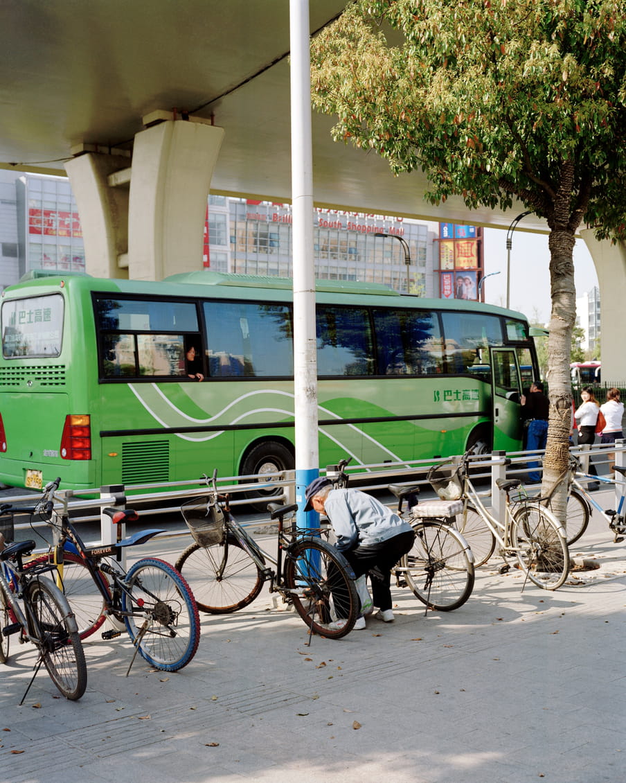 Photograph of a person picking up something from the pavement in between parked bikes. In the background there is a bus and a shopping mall.