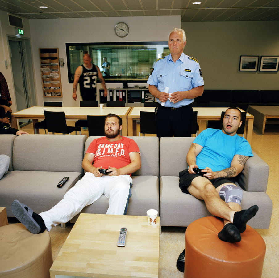 Four people in a photo, two of them in respectively red and light blue t-shirts on a light grey sofa holding black video game controllers, their legs up on soft stools in front of them. A man in prison guard uniform of a blue buttoned shirt and black trousers stands behind the sofa and them, looking in the direction they look in, holding a cup and a rolled up piece of paper. Beyond, a man in a black sports vest walks past two big white tables