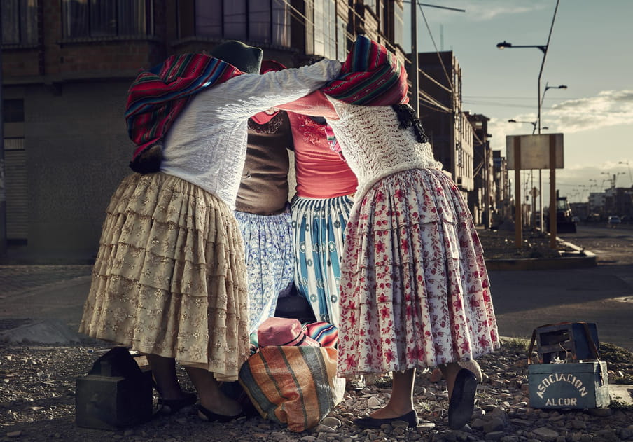 On a sunlit street corner, against a backdrop of blue sky, clouds, a row of buildings, four women in big colourful and patterned, frilly skirts are hugging; the two in front wear white tops and shawls around their backs. We don’t see their heads or faces in the light. In the middle of the huddle is a stripy bag on the floor with a hat in it. There is a small blue box which says ‘Association Alcor’ on it.