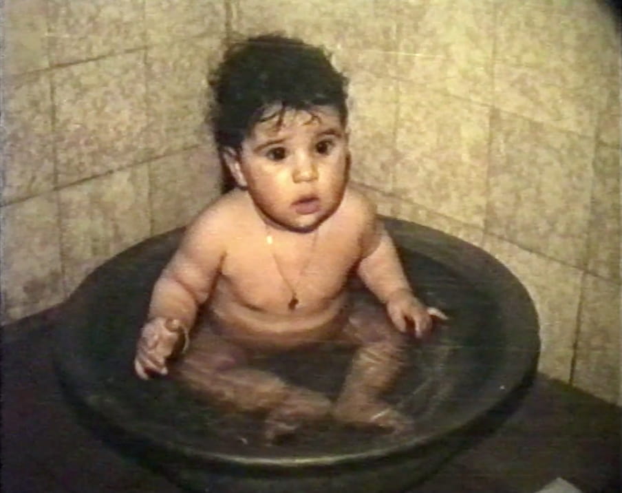 A chubby, naked baby sits in a washbowl in a shower against a backdrop of beige tiles.