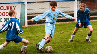 Header image. Photograph of 11-year-old Jamie Lawrence (in the centre of the photo). Three boys playing football in a slightly dated photo; the boy in the centre has arms splayed, and is in a light blue sports long sleeved top and shots, as his right foot is about to kick a muddy football. The other two boys are in darker blue outfits and flank him, eyes on the ball ready to tackle. Behind, we see metallic fencing and a sign saying 'curry'.