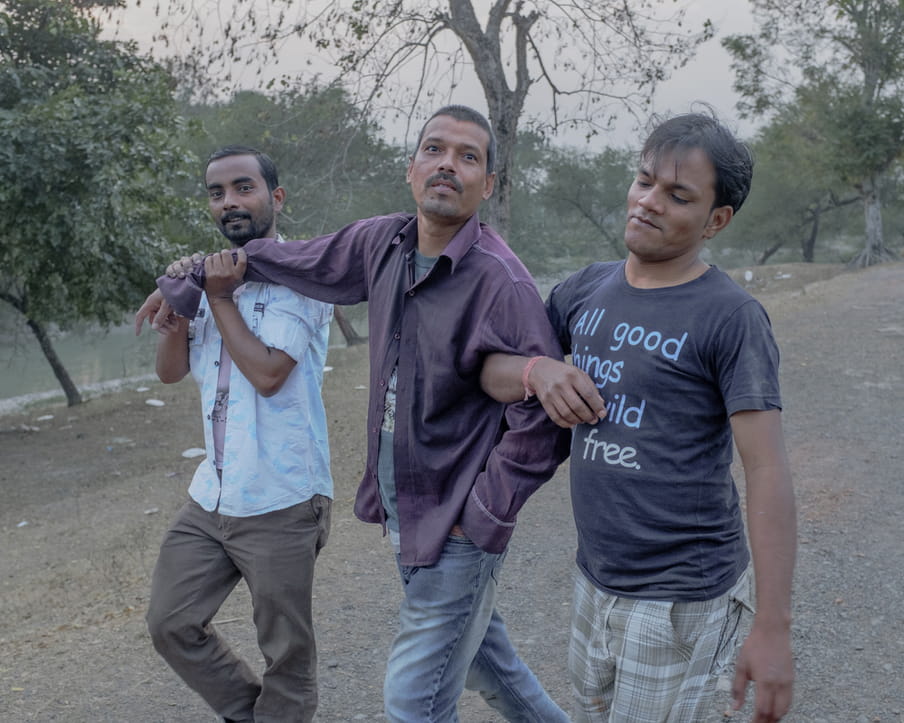 
Three men are walking together on a gravel path. Some water and trees are visible in the background. The one in the middle wears jeans with a purple shirt. He looks at the camera, smiling slightly. The man on the left of the photograph looks at the camera with a small smile while holding the other man right arm at the rist with his two hands. The third man on the right of the frame looks down with a tiny smirk and he is holding the arm of the men in the middle.