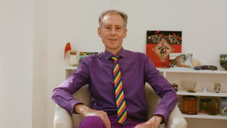 Centre of photo is a person sitting in cream coloured armchair, dressed in purple shirt and trousers, with a stripy red, blue and yellow tie, and with shirt grey hair, hands resting on lap, arms at angles on armrests. Behind is a white bookcase with various objects, including small flags, pots and sunglasses.