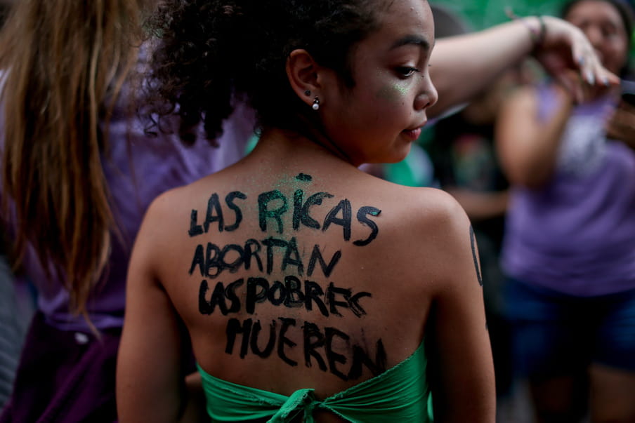 Side profile of a young girl looking to the right in a crowd with the text ‘The rich have abortions, the poor die’ scrawled on her back in Spanish. She has green glitter on her face and wears a green bandana as a strapless top