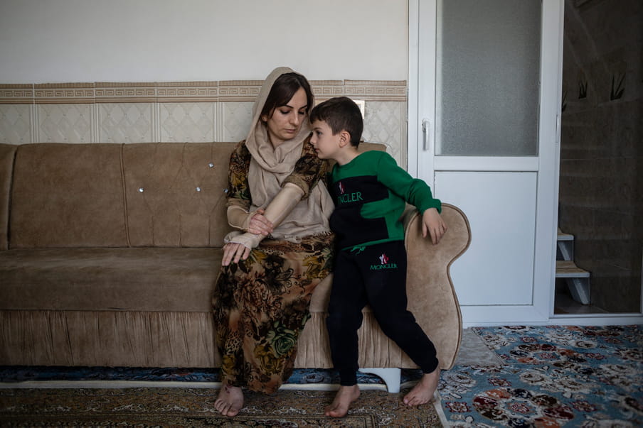 Photograph with two barefoot figures sitting on the corner of a cream velvet sofa. The main figure has a light cream shawl wrapped loosely around their head in a brown dress, and holds a bandaged arm in the other arm. Besides them is a young child in a green jumper at the centre on a couch