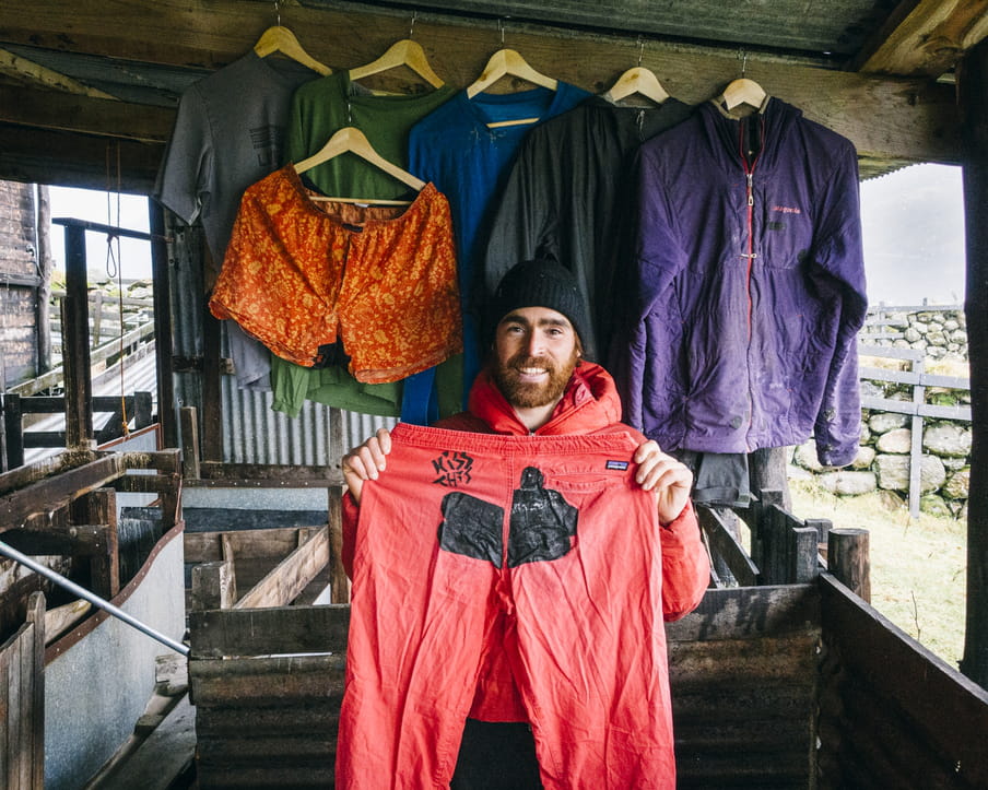 Photo of a man with a beard and a beany, holding a pair of pants up to the camera. In the background several items of clothing are shown.