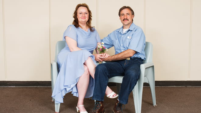 Photo of a man and a woman sitting on plastic chairs in front of a light yellow wall. The woman is wearing a blue dress and has flowers in her hands. The man is wearing hun prison uniform, jeans and a blue shirt. They are holding hands and sitting close together.