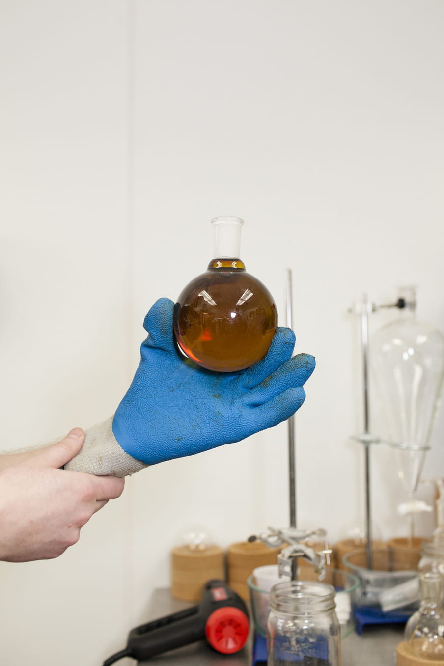 In a close up, one hand holds the other blue gloved hand up - which holds a laboratory bulb shaped flask, almost filled with the brown liquid  of cannabis oil.