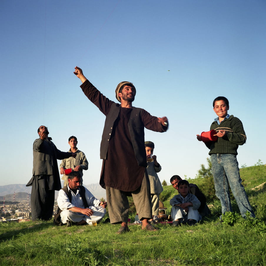 A group of adults and kids standing in the grass with a town in the background. The man in the middle is standing and flying a kite off screen
