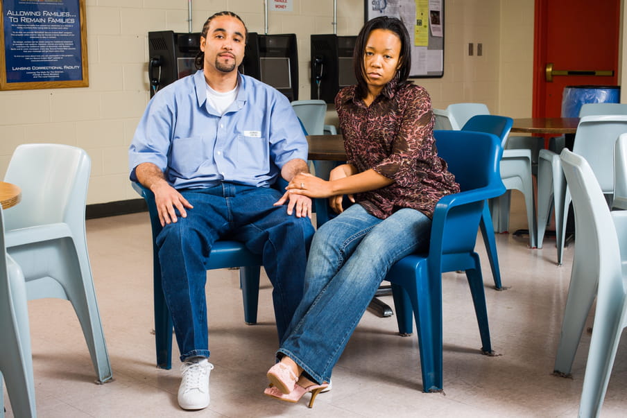 Photo of a man and a woman sitting on dark blue plastic chairs. The woman is wearing jeans and a leopard print shirt. The man is wearing hun prison uniform, jeans and a blue shirt. They are holding hands and sitting close together.