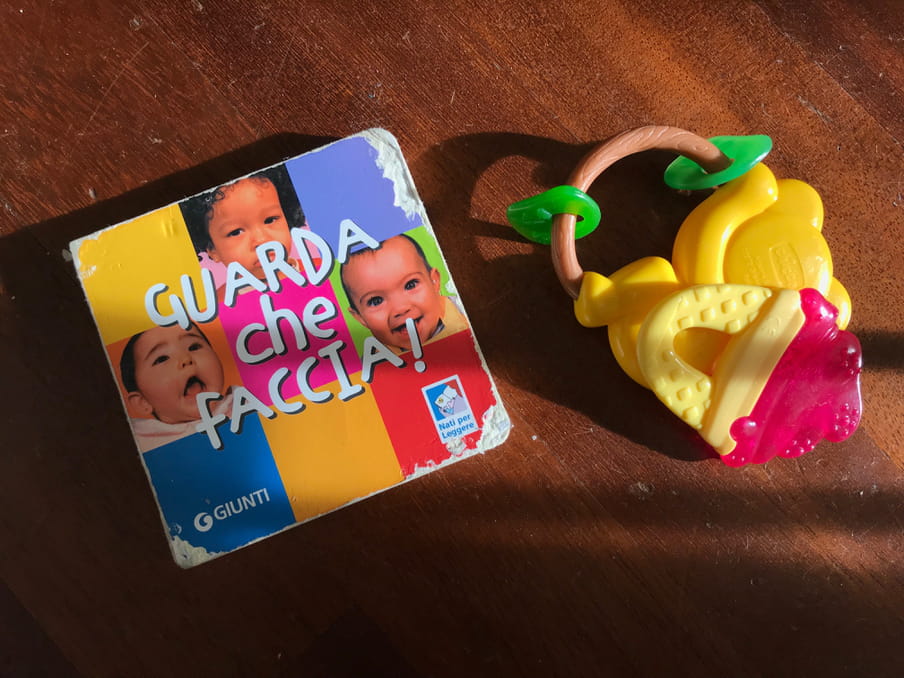 A thick paperboard book in Italian with pictures of babies’ faces is bitten around the corners, and it is on the floor next to two plastic teething toys.