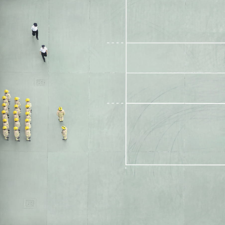 Photograph taken from above on a mint green court showing a group of firemen aligned and two police officers walking towards them.