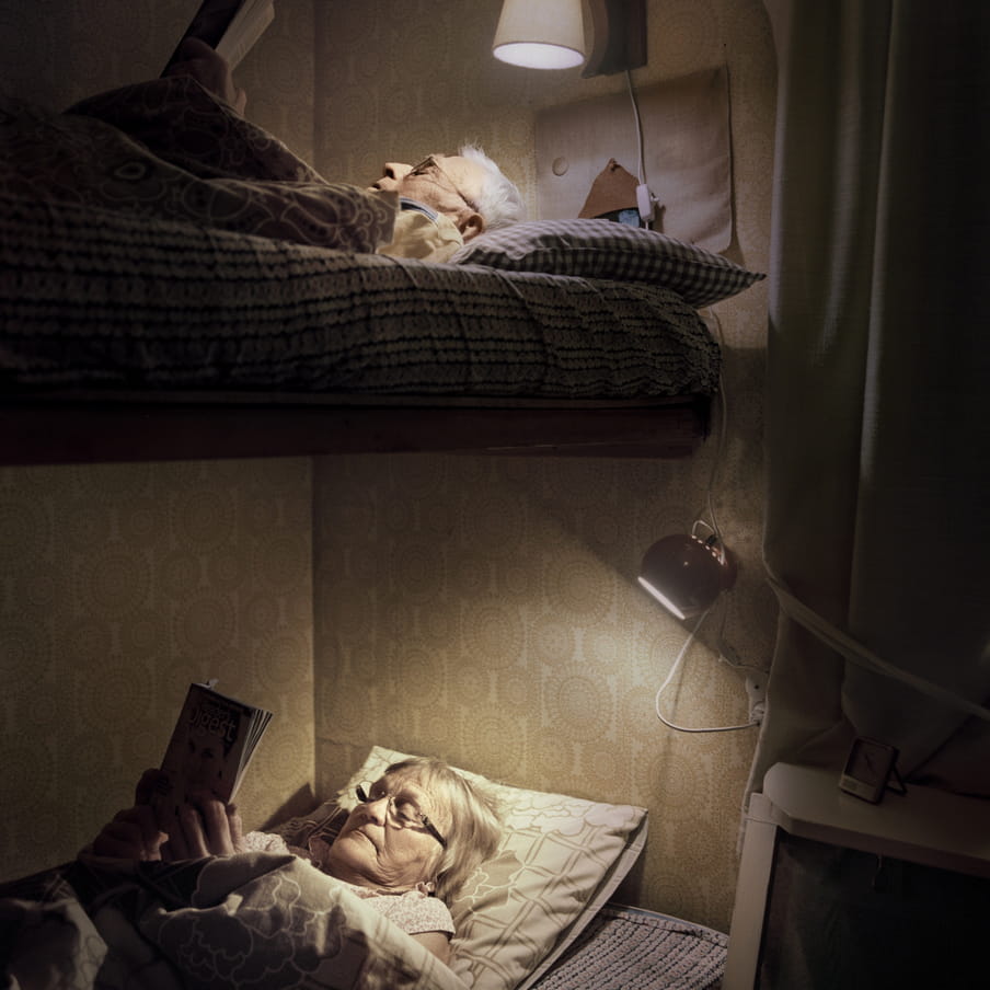 Photograph of an elderly couple reading in a bunk bed. Two small bed lights shine on the faces. 