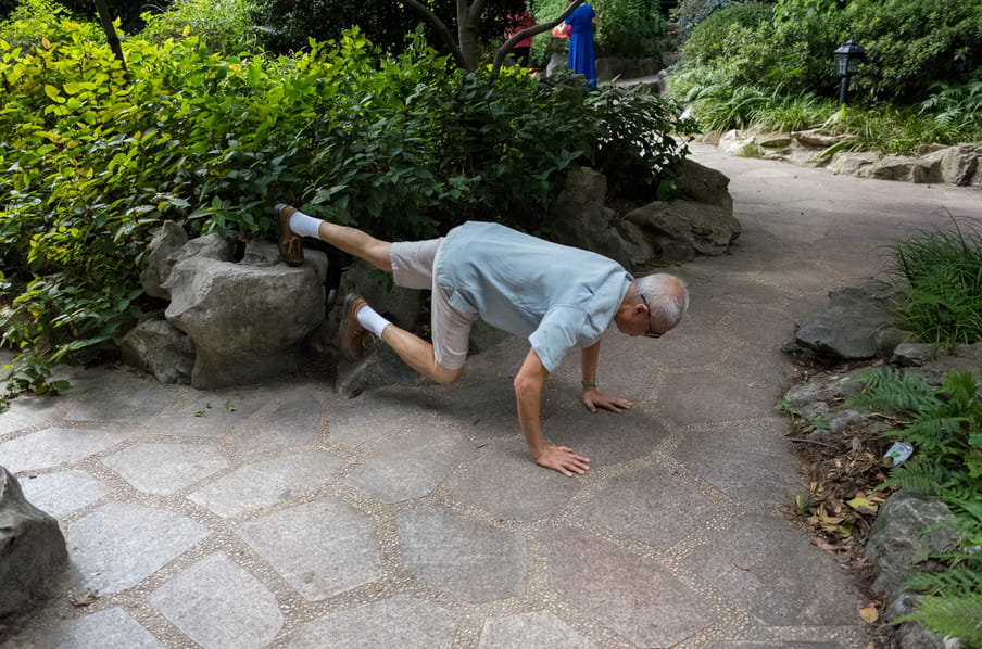 And old man in a park, leaning over a stone path to do stretching exercises.