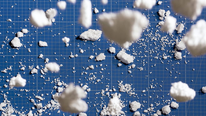 Photograph of white pieces of styrofoam looking like little clouds on a blue gridded background; a few of them are close up the camera, blurry clouds, while the rest lie in smaller pieces in the background, giving us a sense of dimension