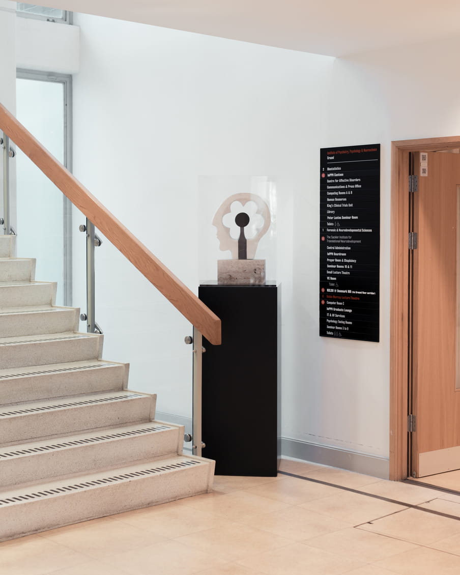 Hallway in a university with on the left a staircase, on the right a door with a sign next to it describing the different departments and their floor numbers. In the middle stands a statue of a head, with a flower like icon where the brain would be.