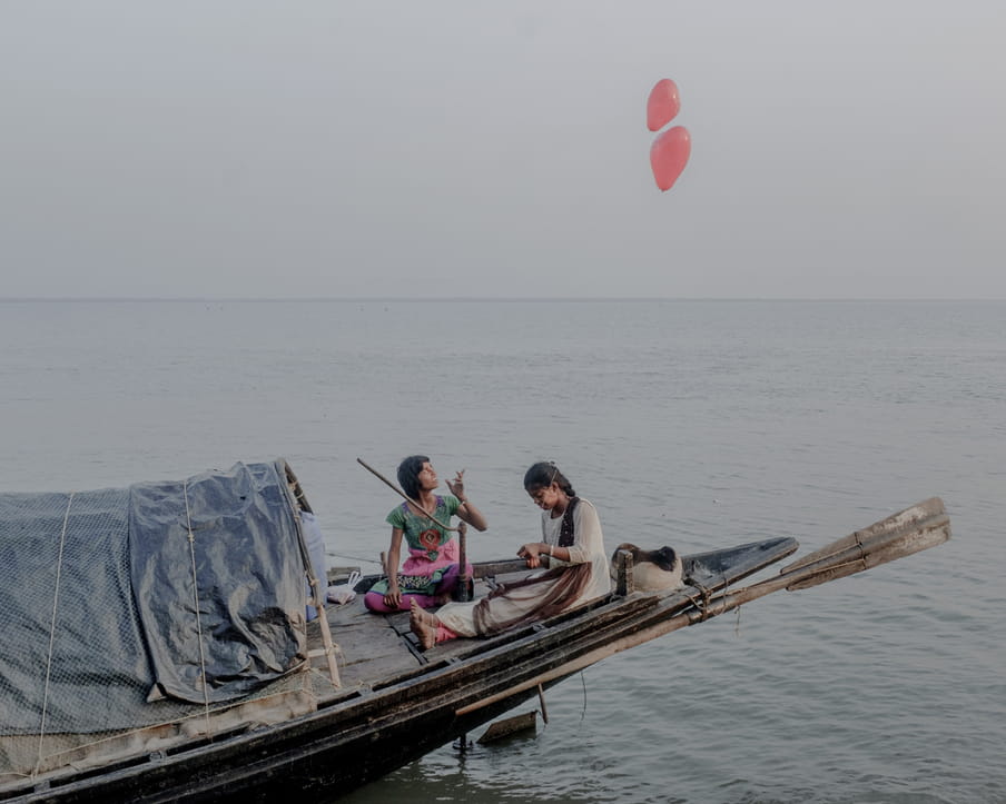 Two women sit on the bow of a long fishing boat on the water, smiling, one with a plait, in white clothes, and with something in her hands which she looks at, her legs crossed at her bare ankles and feet. The oar is at rest behind her. There is tarpaulin covering the boat’s hull. The other woman, in a simple green, pink and purple shift, seems to have just left two heart-shaped red balloons into the air, her hand poised as she looks up at the sky. We see the horizon in the background. 