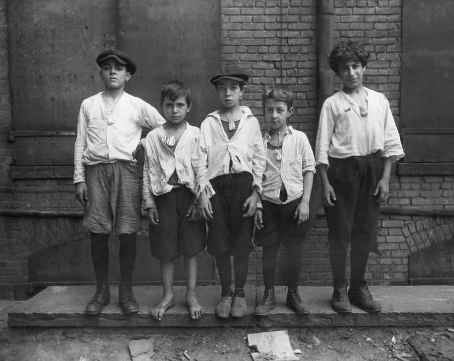 Black and white photograph of five boys standing outside wearing a small bag around their neck.