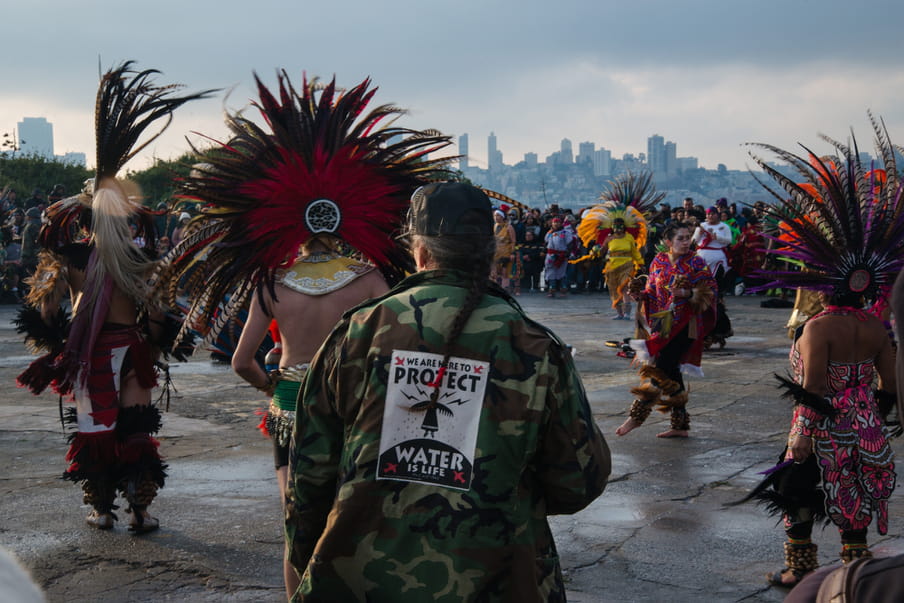 Photo of people dancing in traditional attire and a man wearing a jacking with the text ‘We are here to protect - water is life’