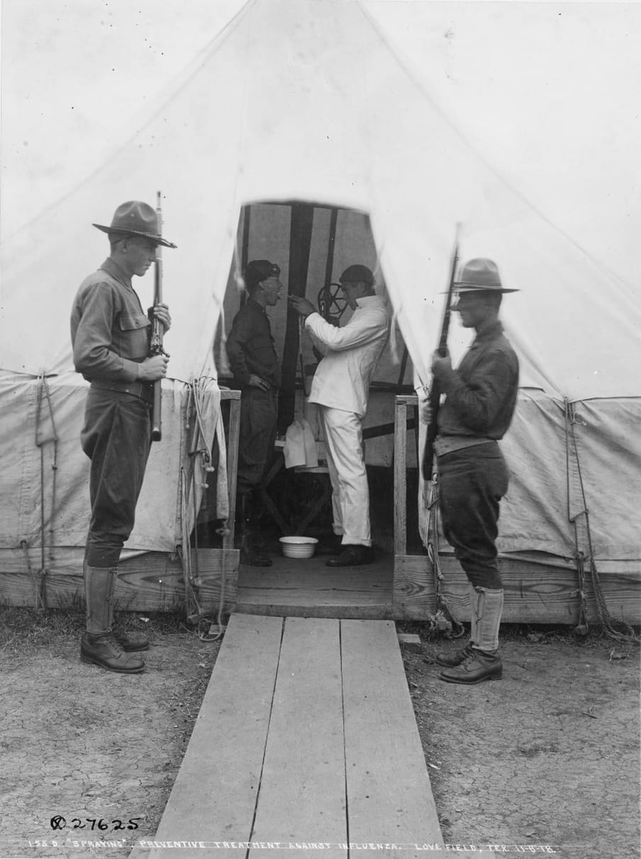 Black and white photograph of a tent, gard by two soldiers while inside a man is putting an object in another soldiers mouth.