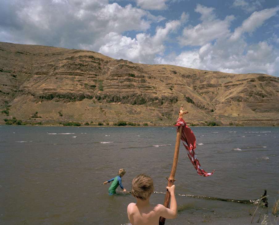 Photo of two kids playing in a river, one has a branch with a flag-like thing on it. Mountains are in the back.