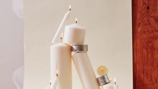 Beige lit candles of various sizes are stack together, in balance. The background is also beige with a woody surface visible. 