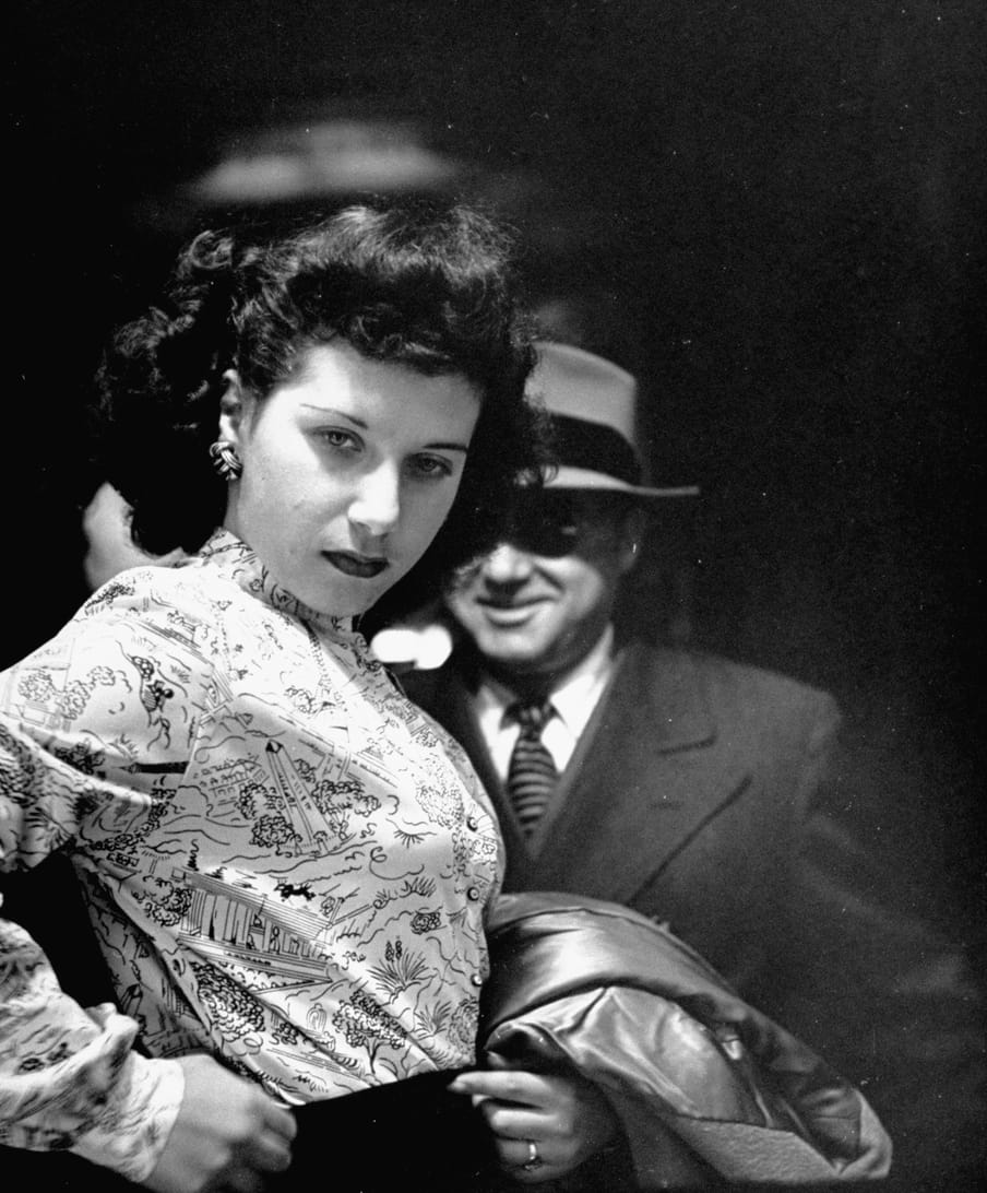 Old black and white photo of a woman checking her skirt while watching herself in a mirror, photographed from the back of the mirror. A man stands behind her