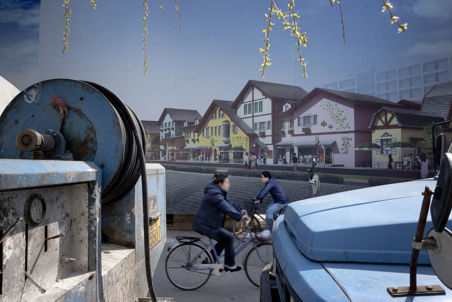 Two people cycling in opposite directions in front of a wall showing a photograph of a peaceful strip of houses.