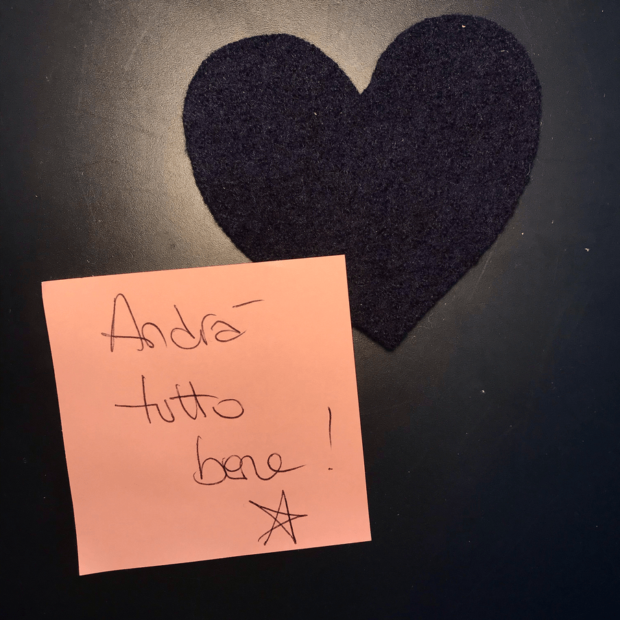 The image shows a dark blue felt hear and a handwritten pink post-it note that says: Andrà tutto bene.