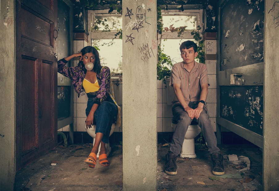 Screenshot from the series Sex Education showing a boy and a girl sitting in abandoned toilet stalls, the girl blowing bubblegum. The boy looks uncomfortable.