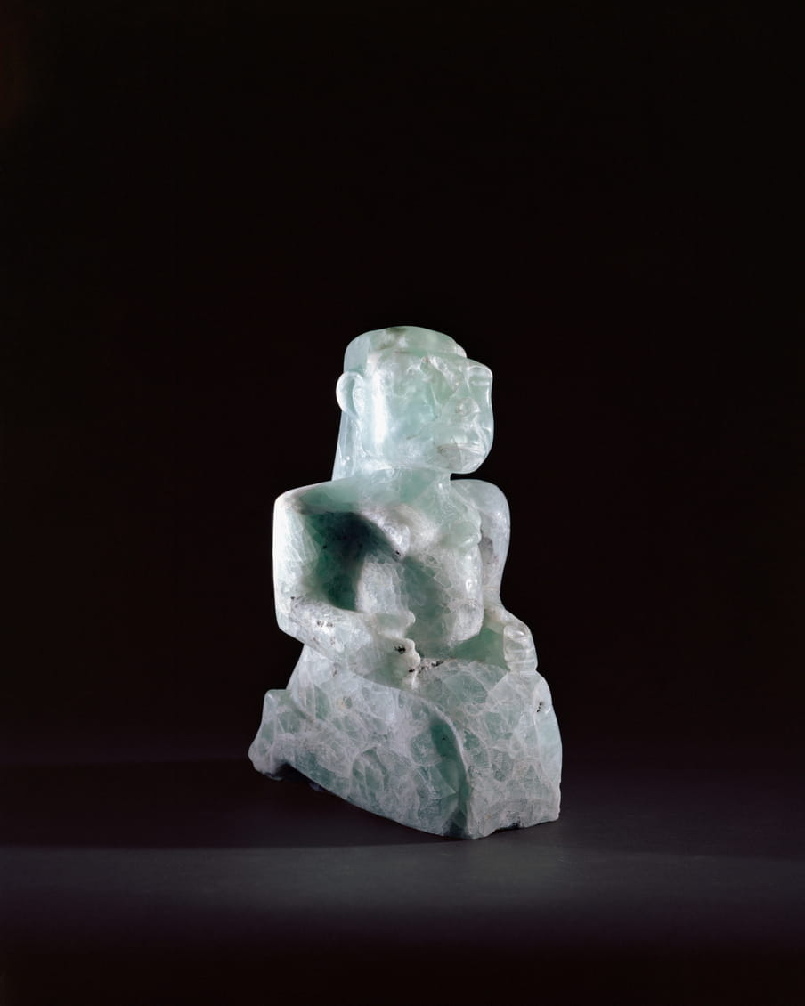 Photo of a stone statue of a figure sitting up straight on its knees - on a black background. It’s much less smooth and refined that the one in the last image, and no cape is visible.