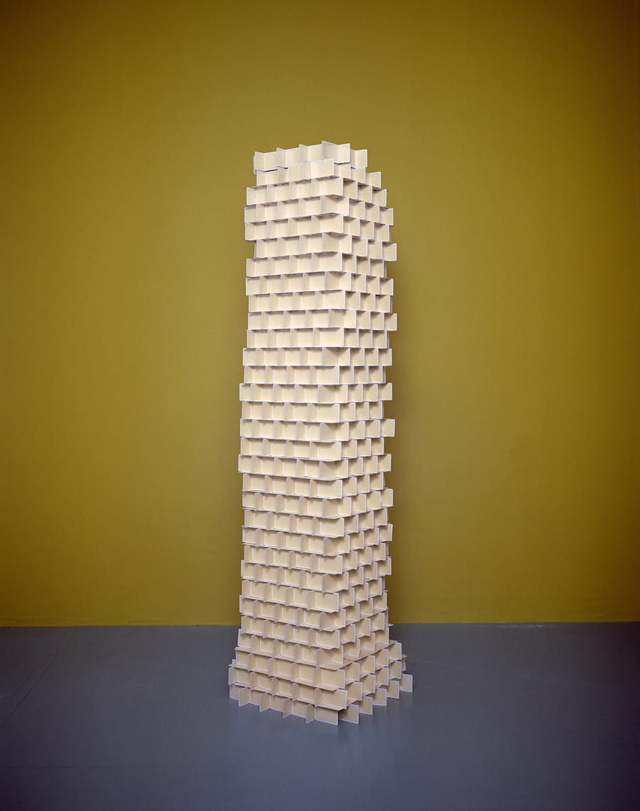 A huge column made of yellow playing cards stands in front of an ocher wall and lays on a grey floor. 