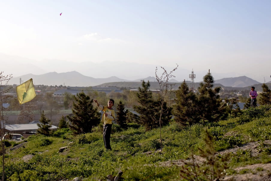 Two boys, one in yellow sweatshirt and one in a pink one are flying kites, with hills and a town in the far distance.
