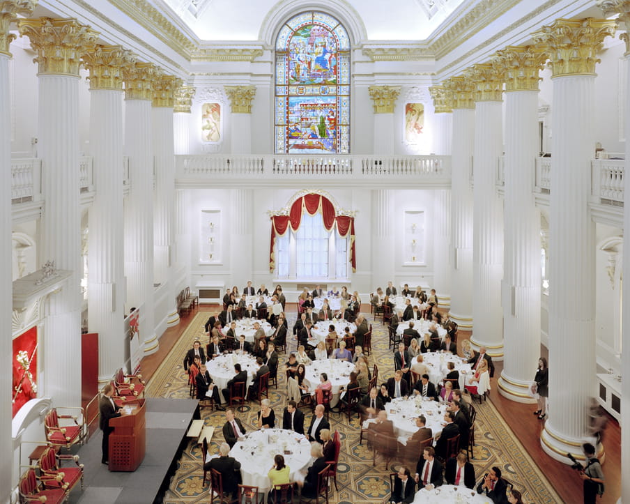 Picture of a large hall with roman white and gold columns, and about twelve round tables with white table cloths, people around it, looking at one man on a stage speaking