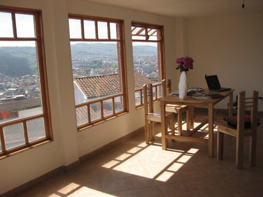 A photograph of the correspondent’s apartment in Quito showing a table with four chairs and a vase with flowers, and on the left the picture window with a view of the outside rooftops.