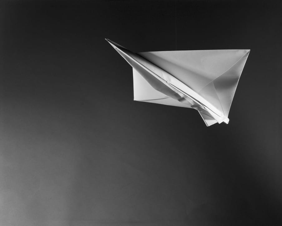 Black and white photo of a white paper plane, nose up in flight, to the top right of the image