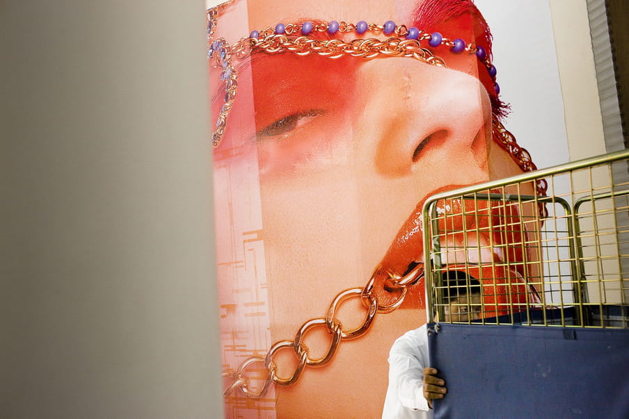 A man in white is pushing a high cart in front of a huge poster of a women’s face with an intense make up and necklaces around her mouth, eyes and forehead.