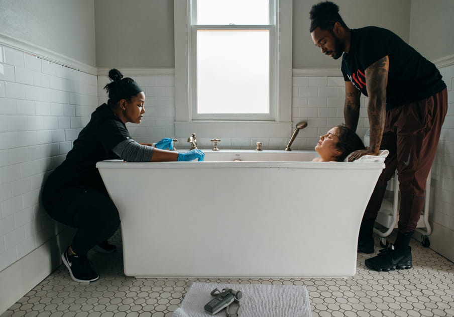A narrow bathroom with circular floor tiles. A woman with bright blue latex gloves kneels at one end of the bath in black outfit and trainers and with a top knot and a headband, tending to a birth; a man stands with hands on a towel pillow at the other end of the bath, in black boots, red trousers, a black t-shirt, and with hair in a bun. A woman lies in the bath, her head resting, eyes closed. There is a bright window behind the scene, and a paper towel laid out in front of the bath, with silver tools.