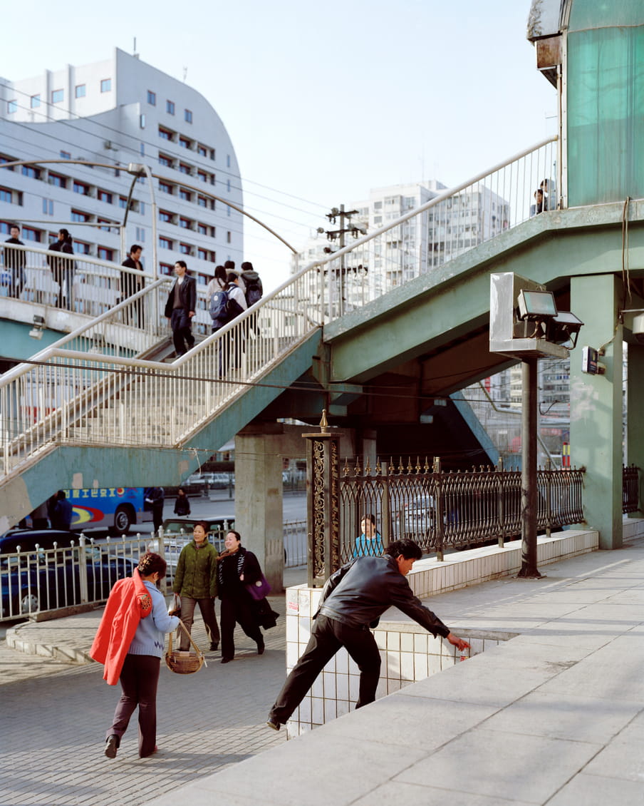 Photograph of a person picking up something that looks like a bottle from stairs. In the background there are people walking on the pavement and on multiple stairs going over the road.