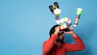Man in front of a blue background - looking trough self-built binoculars, made from duck tape, toilet rolls, plastic and cups. It is warped and too long.