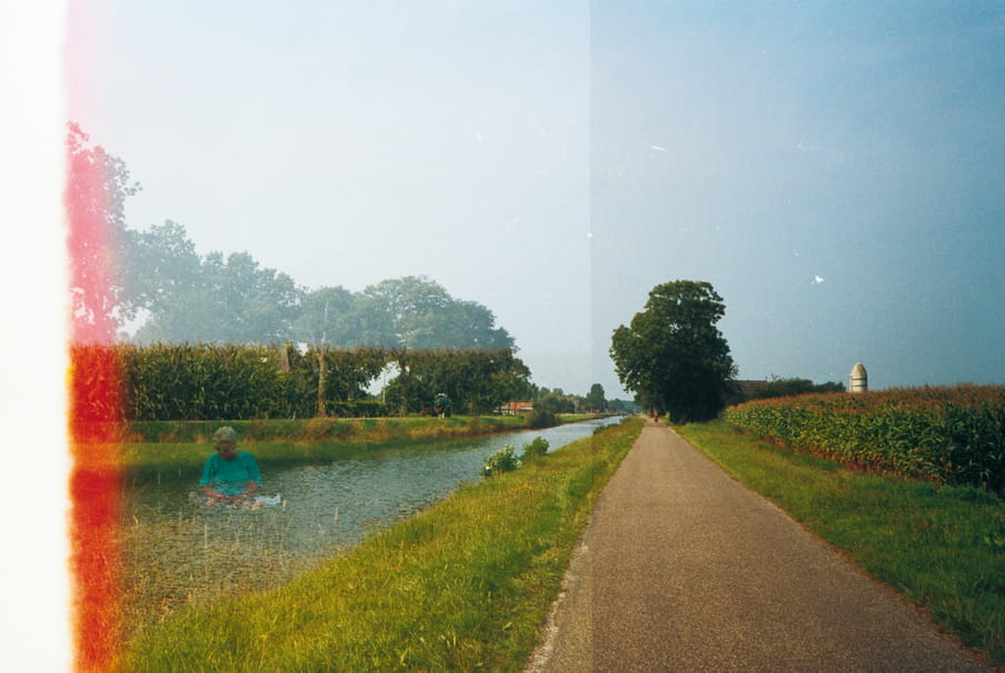 Photo of a road with a river next to it. Due to double exposure, a man seemingly sits in the middle of the river. A light flare cuts of the image on the left side.