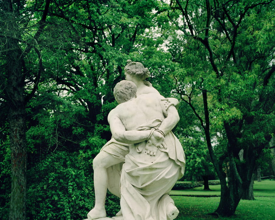 A classical statue of two people, their fingers are broken off, in a green garden full of trees.