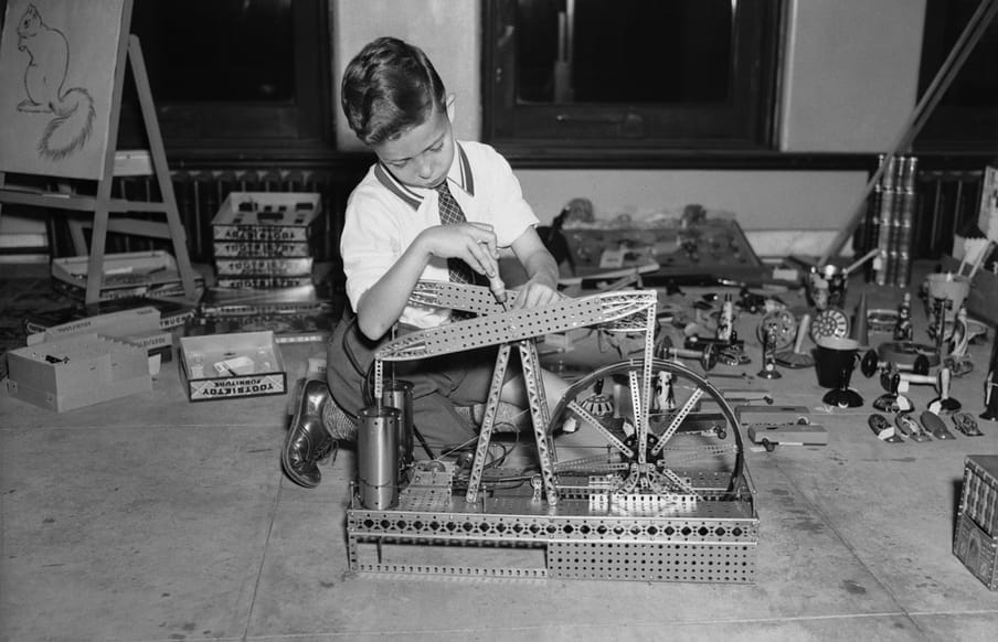 Old black and white photo of a boy, wearing formal clothes building a metal spinning wheel