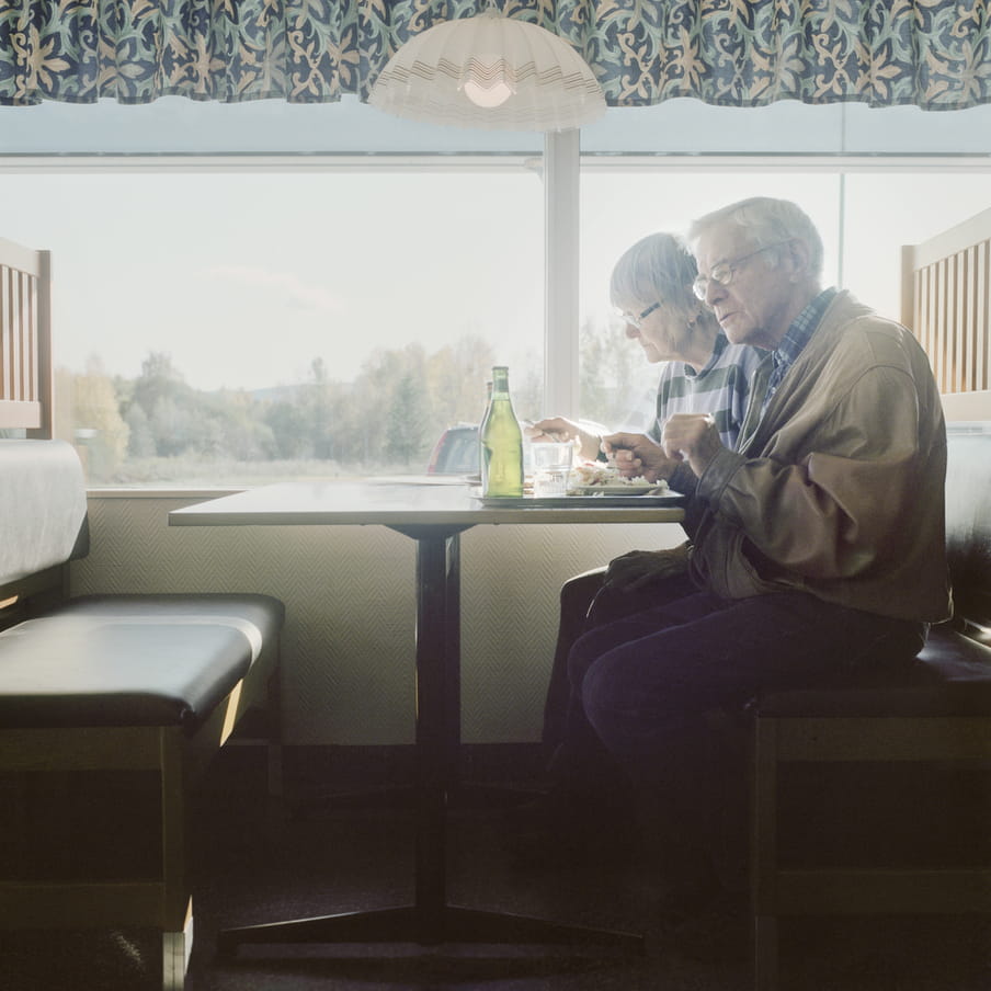 Elderly couple sits in a diner booth in front of a window that shows a green scenery. On the table there is a tray with food and drinks. 