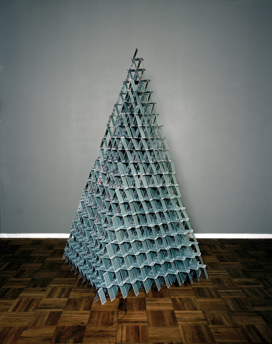 A huge pyramid made with turquoise playing cards lay on a parquetry floor. The back wall is grey with a white baseboard.