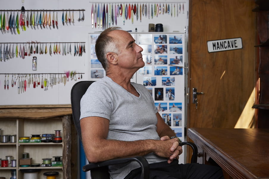 Photo of a man sitting in a room in a grey shirt - in the back fishing equipment and photos of him with fish